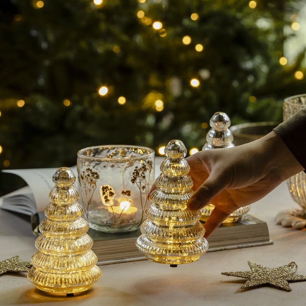 Aurora silver tree portable lamps glow warmly on a festive table as a hand adjusts one tiered lamp beside a candleholder and scattered stars.