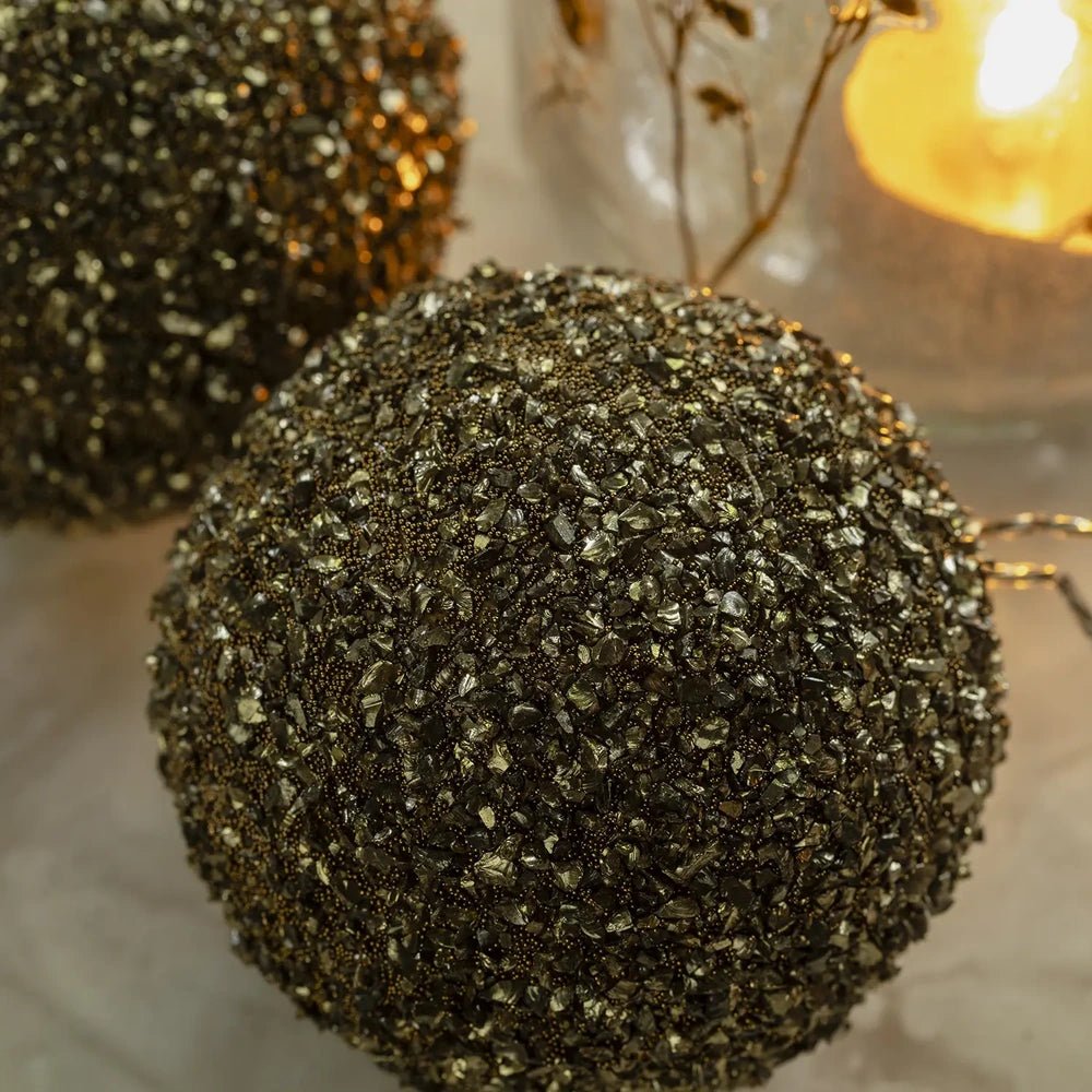 Ayla beaded bauble shows a densely textured round surface covered with dark gold beads, resting beside a glowing patterned candleholder on a festive table.