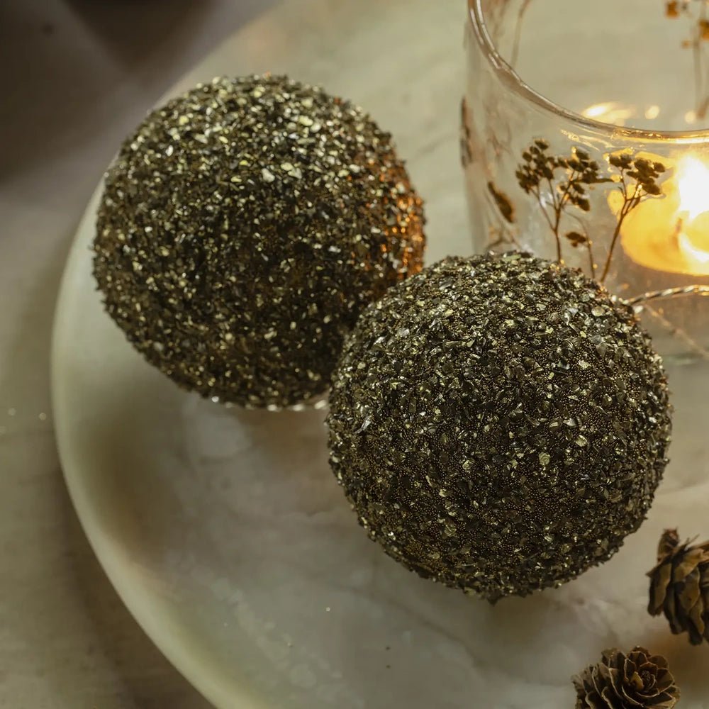 Ayla beaded baubles in two sizes sit together on a pale tabletop, showing their densely textured gold surfaces beside a glowing patterned glass candleholder.
