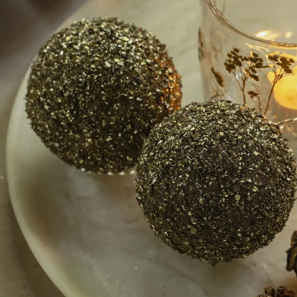 Ayla beaded baubles form a pair of textured gold spheres beside a patterned candleholder, with warm light highlighting their clustered bead surfaces on the table.