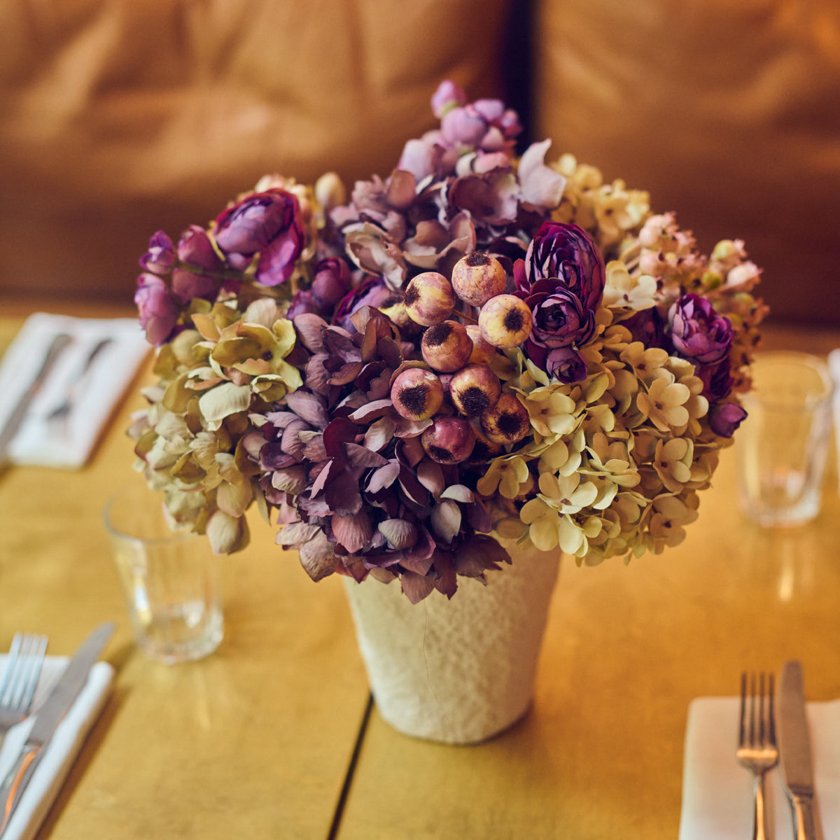 Bouquet of faux flowers in a vase on a wooden table with cutlery and glasses.