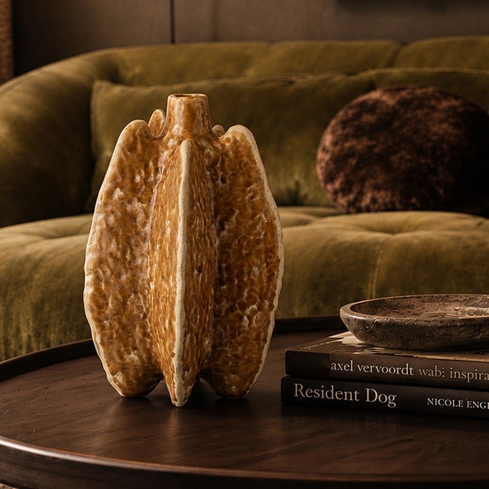 Bexley Porcelain Vase with sculptural folded form and warm ochre finish, styled on a dark tray beside stacked books and an olive green sofa.