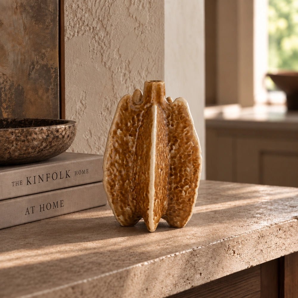 Bexley Porcelain Vase with sculptural folded form and warm ochre finish, displayed on a pale stone console beside stacked books in a softly lit neutral interior.