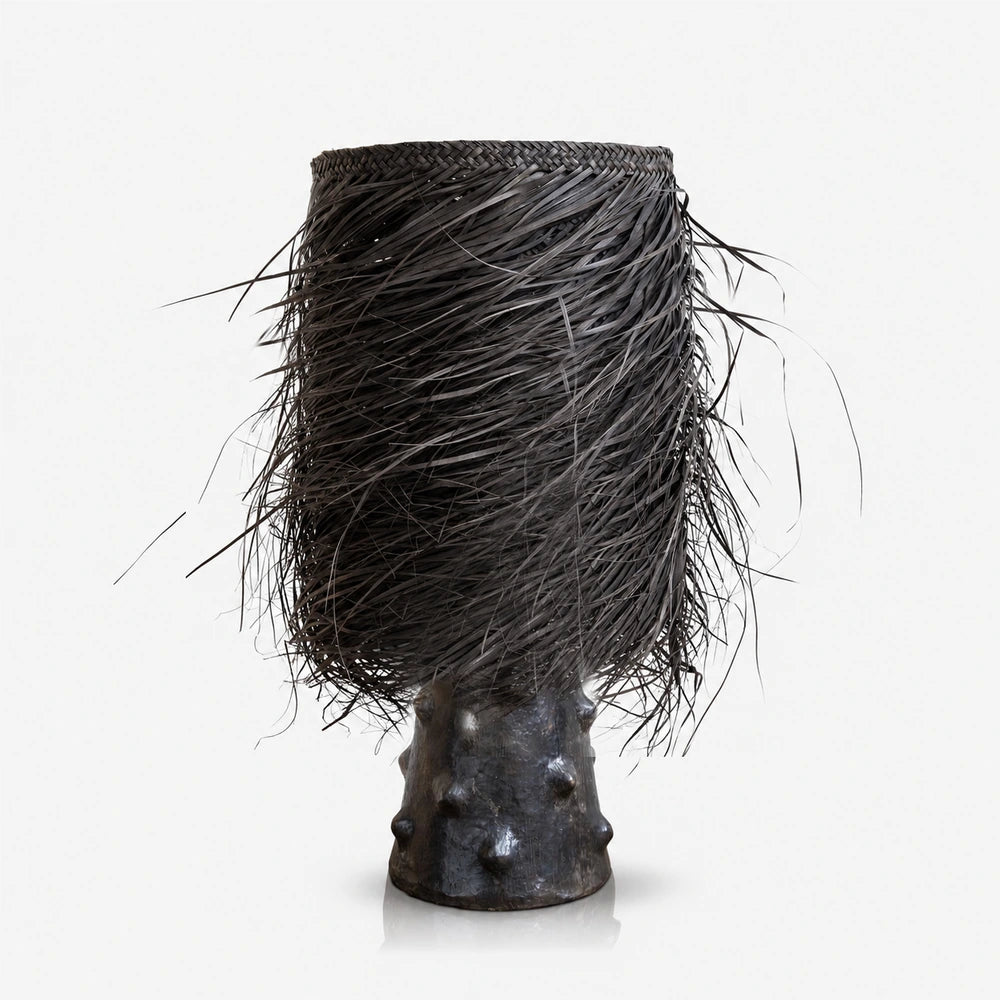 The Black Shaggy Palm Lamp features a tall cylindrical woven shade with loose black fringes and a dark sculptural base with raised textured details.