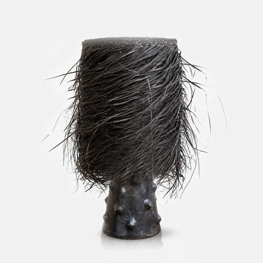 The Black Shaggy Palm Lamp features a tall cylindrical woven shade with loose black fringes and a dark sculptural base with raised textured details.