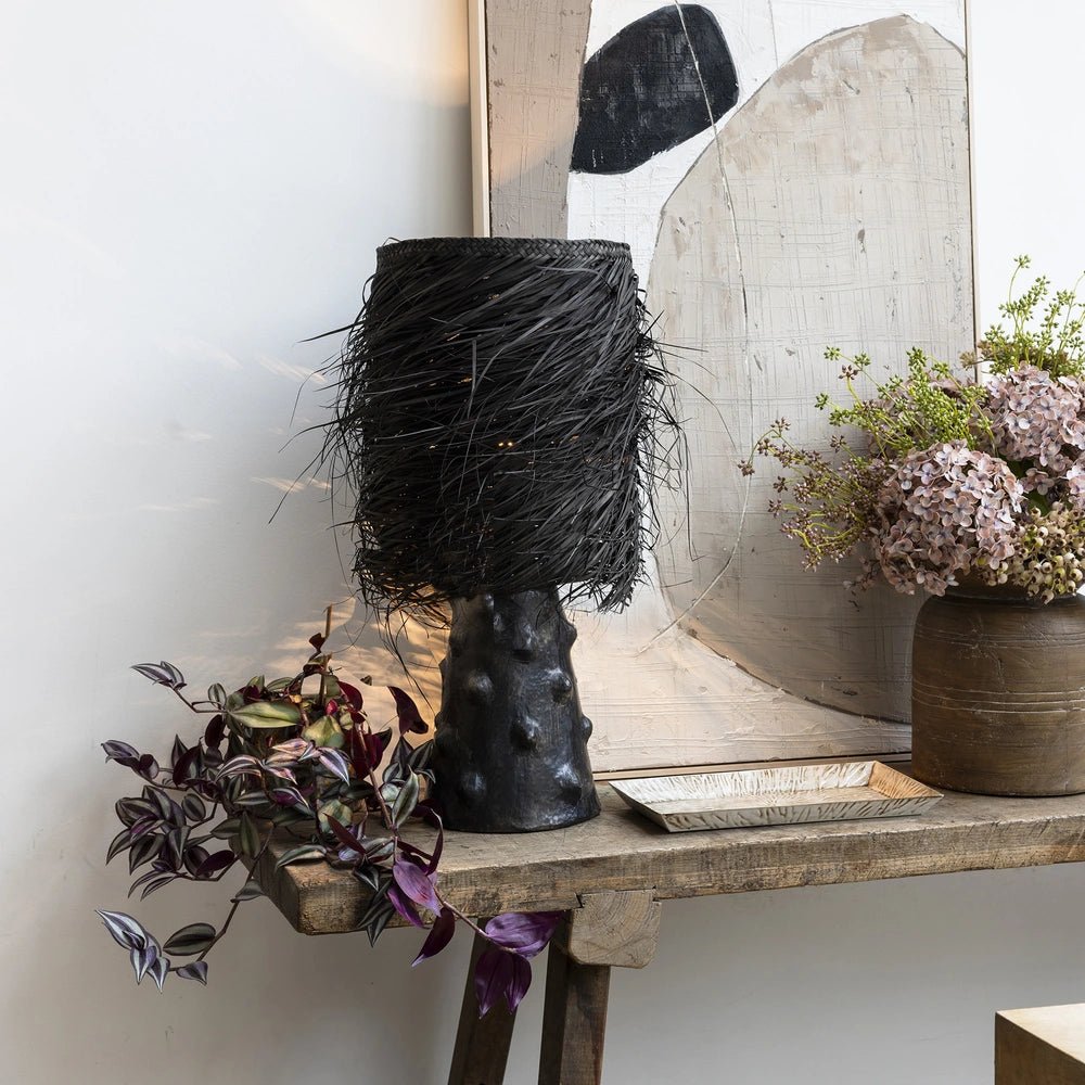 The Black Shaggy Palm Lamp features a fringed woven shade and sculptural dark base, glowing on a wooden console beside trailing foliage and pink flowers.