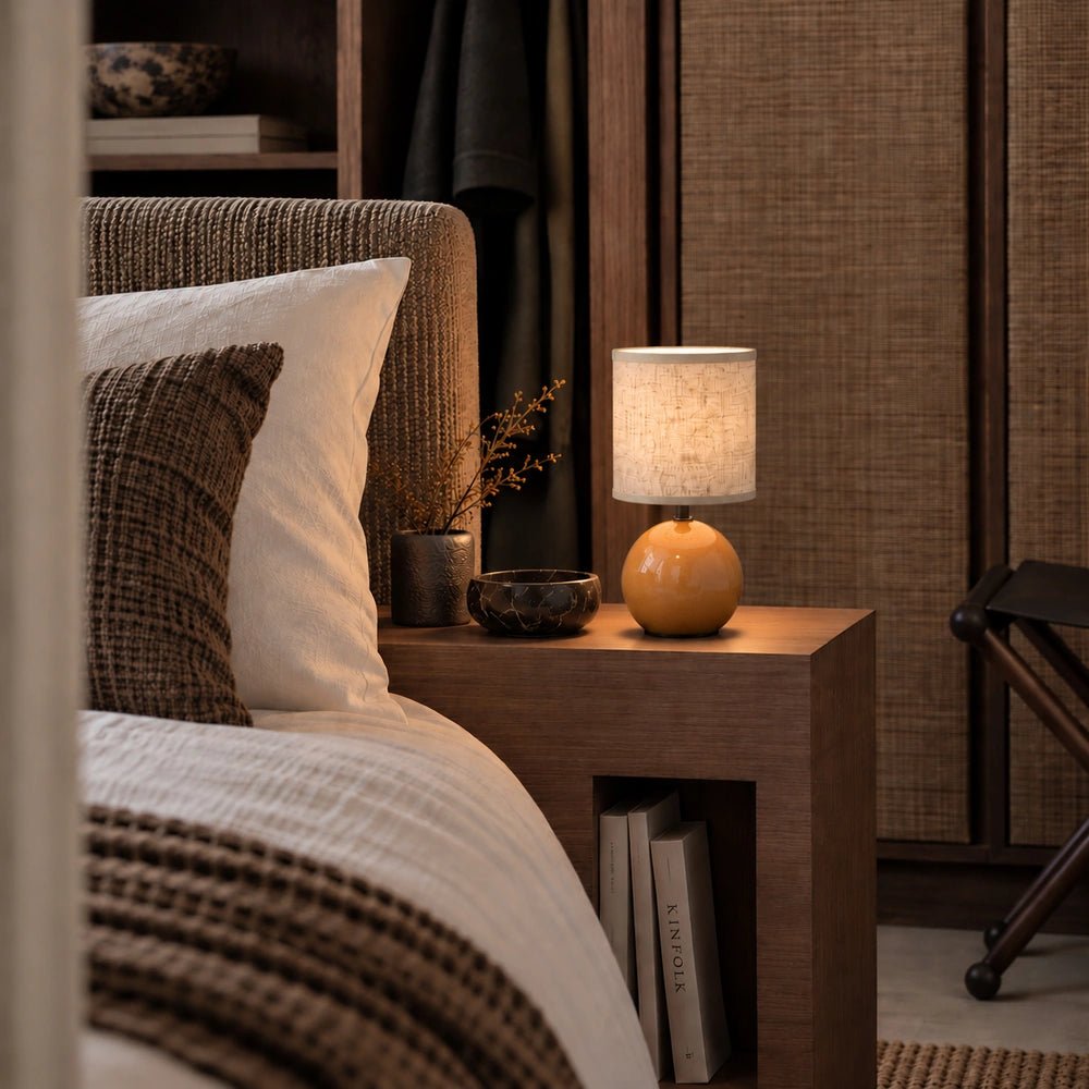 Orange Blair Table Lamp with rounded glossy base and fabric shade glowing on a wooden bedside table beside layered neutral bedding.