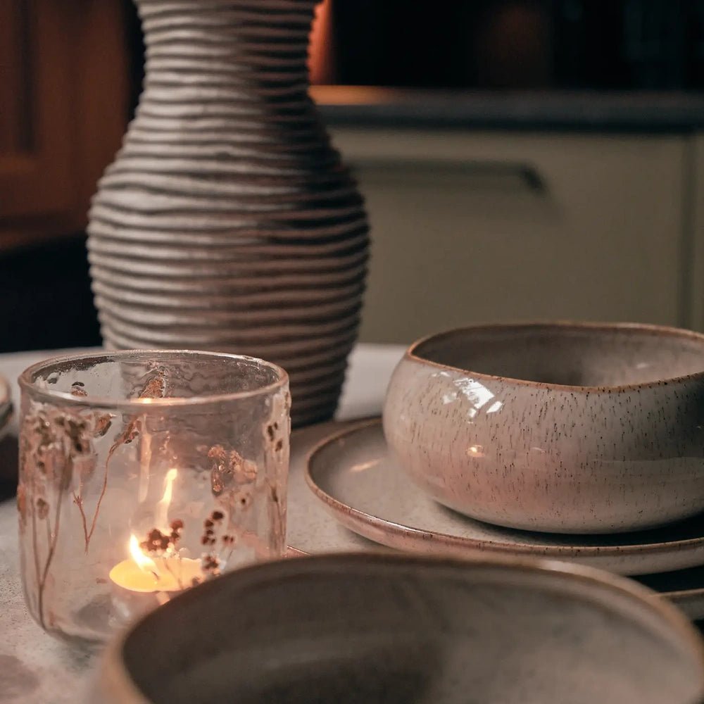 Botanic Glass Candle Holder in medium size with a mottled translucent surface, glowing on a dining table beside a ribbed vase, bowl and plates.