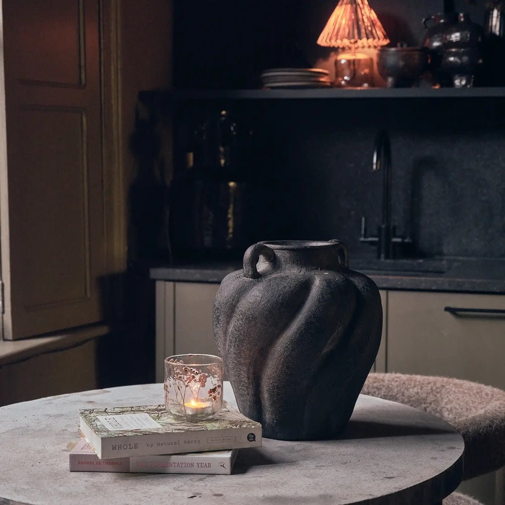 Botanic Glass Candle Holder in medium size with a translucent mottled surface, glowing on stacked books beside a dark handled vase on a round table.