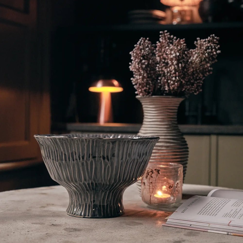 Bowham Ceramic Decorative Bowl with a dark ridged finish and flared shape, styled beside a candle and a vase of dried stems on a table.