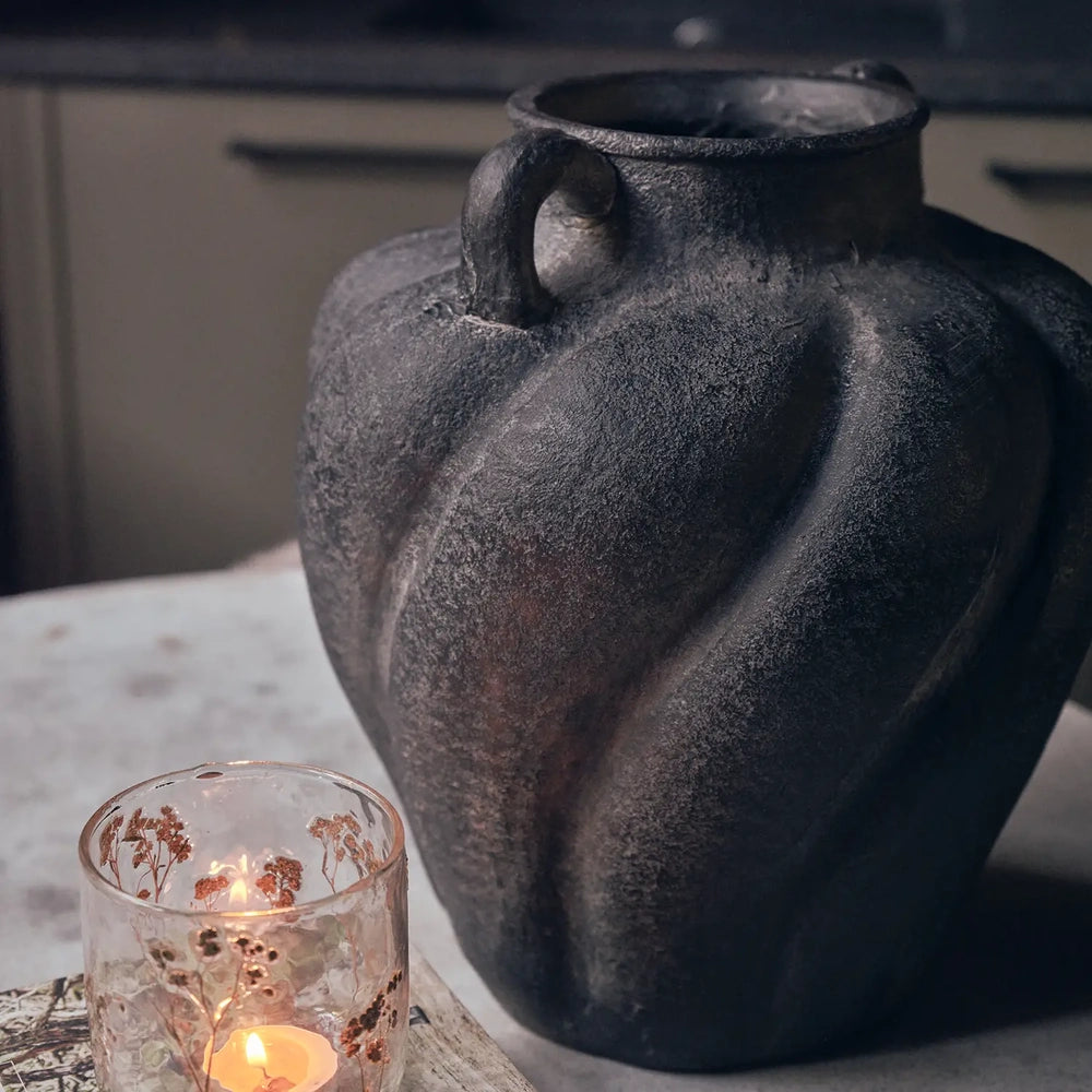 Braddock Magnesium Vase with a dark textured finish, rounded body and small handles, displayed beside a glowing glass candle on a pale tabletop.