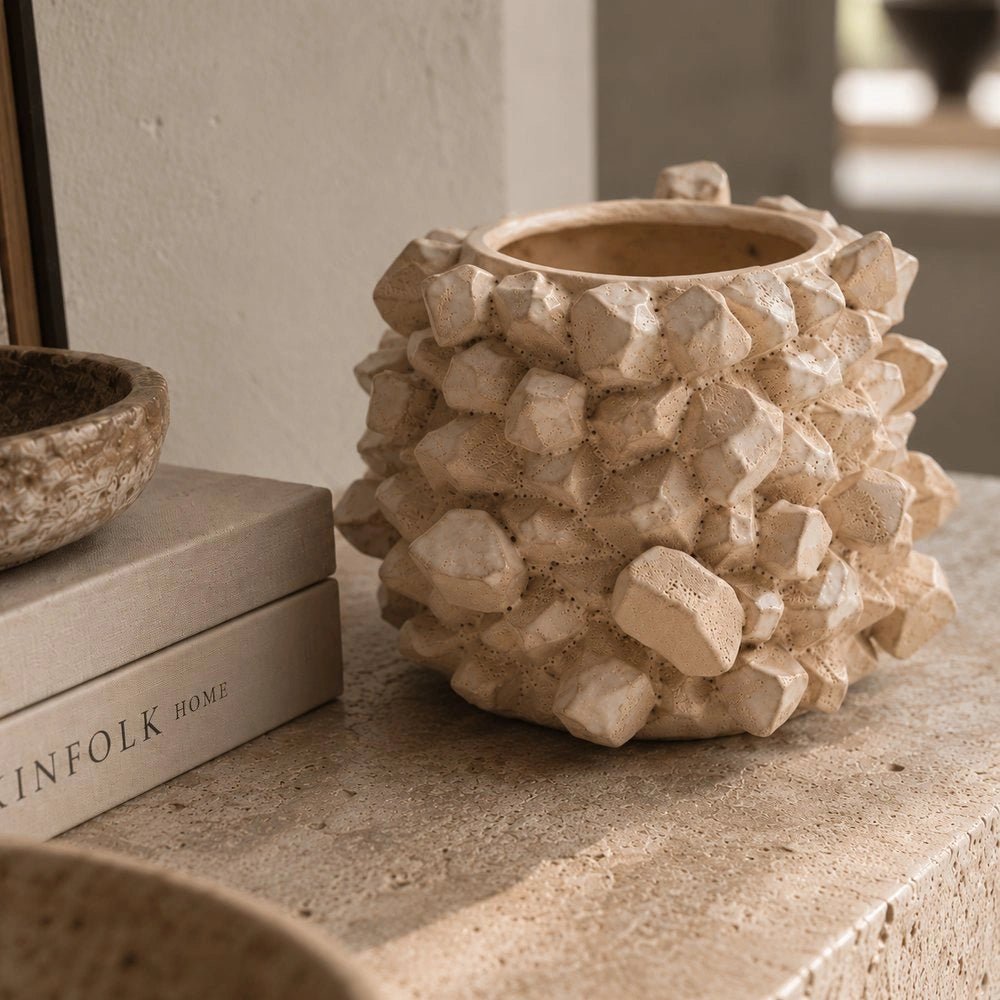 Bricket Textured Vase with a pale heavily textured faceted surface, styled on a console beside stacked books, neutral ceramics and softly lit interior decor.