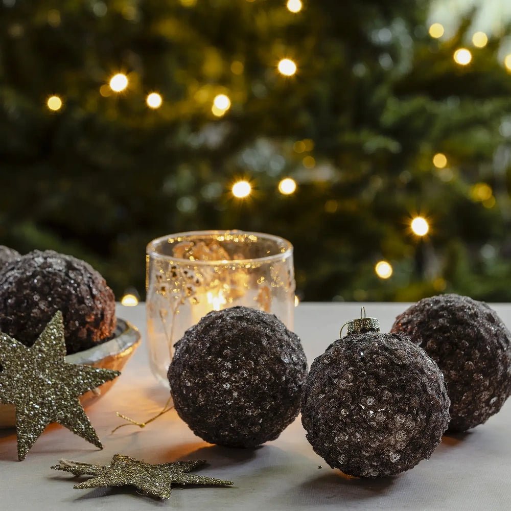 Brown Sugar sequin baubles in dark brown tones rest beside a patterned candleholder, with clustered sequins catching warm light against blurred Christmas tree lights.