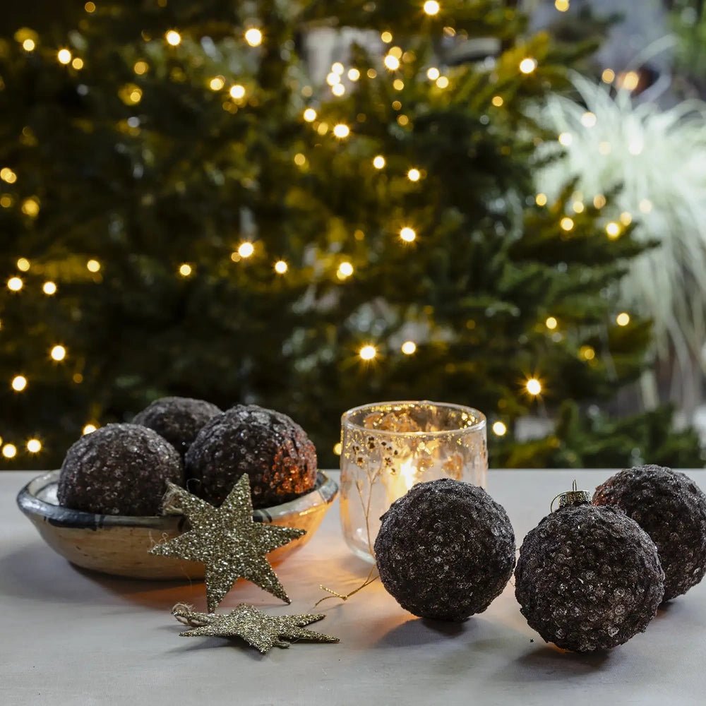 Brown Sugar sequin baubles with rounded shapes and dark textured surfaces sit beside a glowing candleholder, decorative stars and a bowl on a festive table.