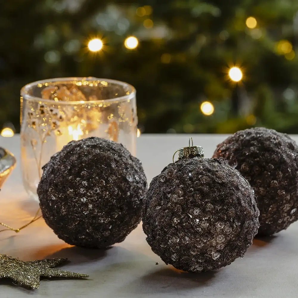 Brown Sugar sequin baubles display densely layered dark brown surfaces in close view, arranged beside a glowing patterned candleholder on a pale festive tabletop.