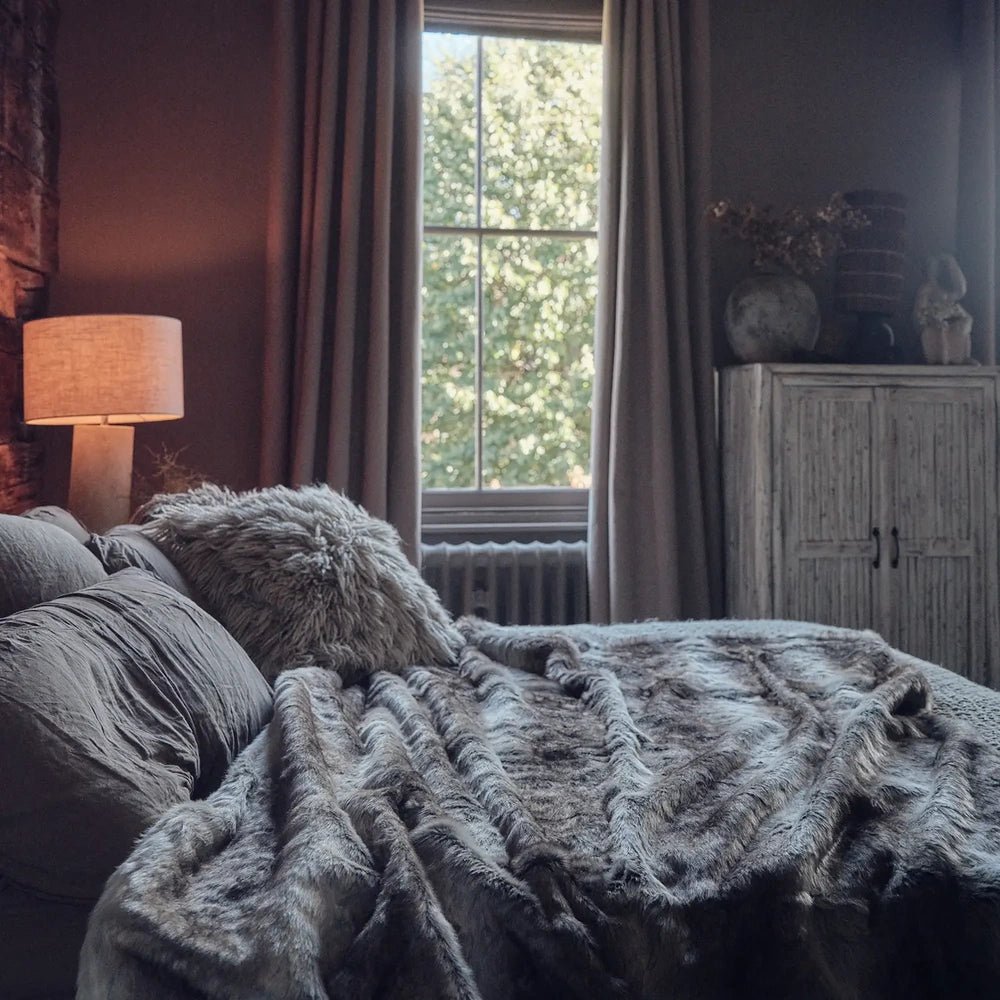 Calvert Faux Fur Throw with deep grey textured folds, spread over a sofa beneath a window beside a softly glowing lamp and dark cabinet.
