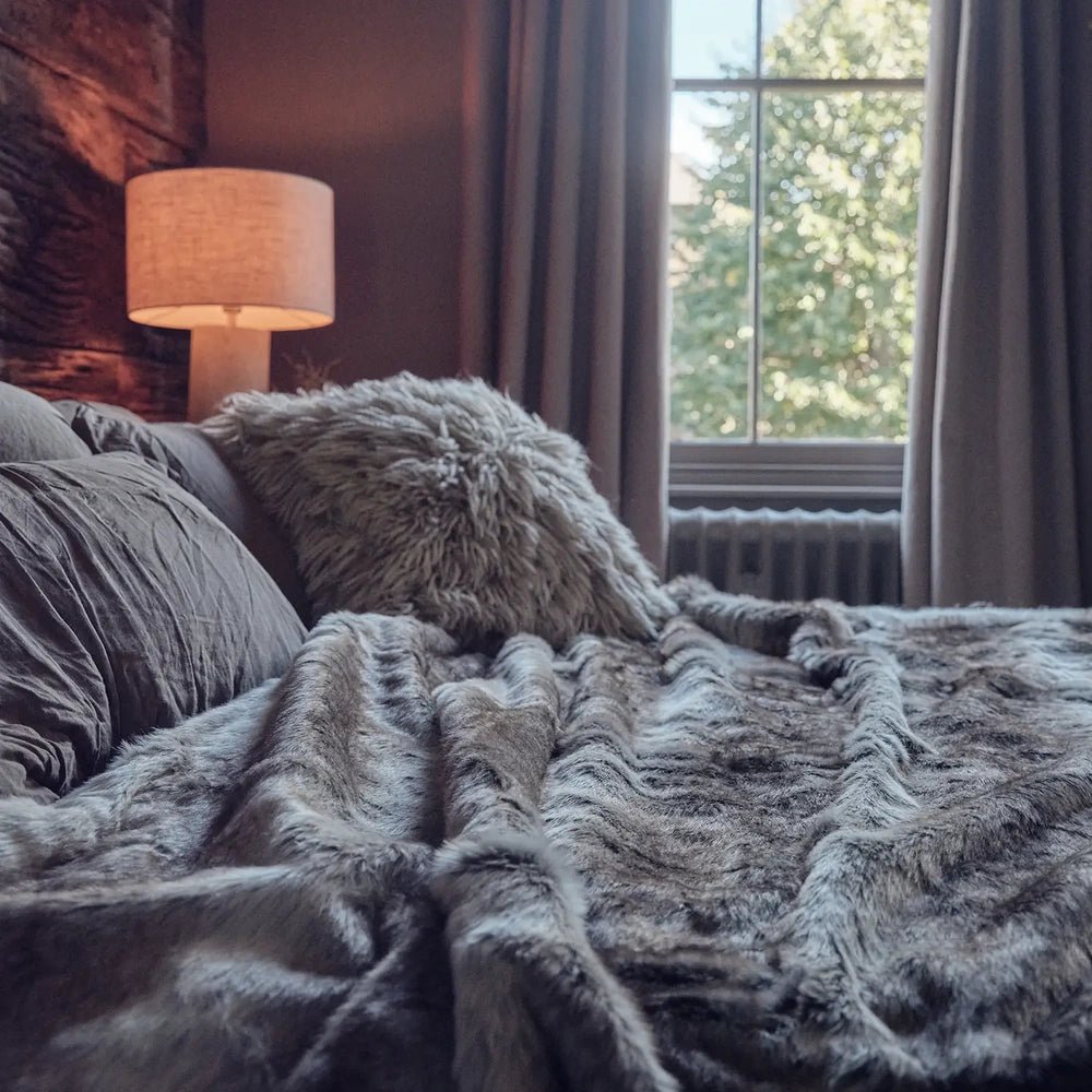 Calvert Faux Fur Throw in grey, draped across a cushioned sofa beside a window and warm table lamp in a dark, cosy living room.