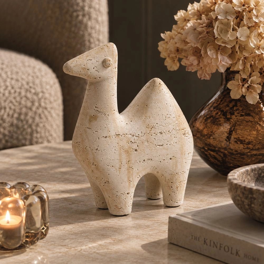 Camel Abstract Sculpture displayed on a dark side table, showing its pale textured animal form beside stacked books, a lamp and neutral decorative objects.