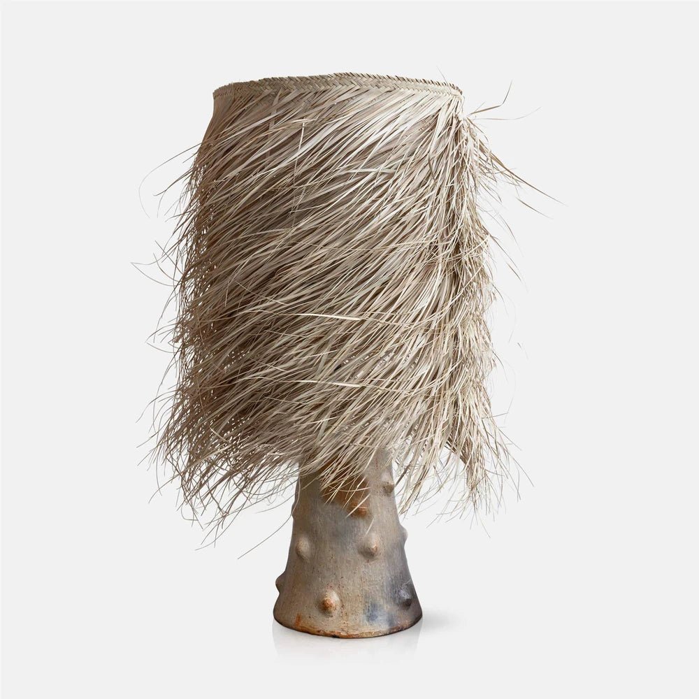 The Caramel Shaggy Palm Lamp has a tall pale beige woven shade with loose fringed strands above a tapered sculptural base with raised textured details.
