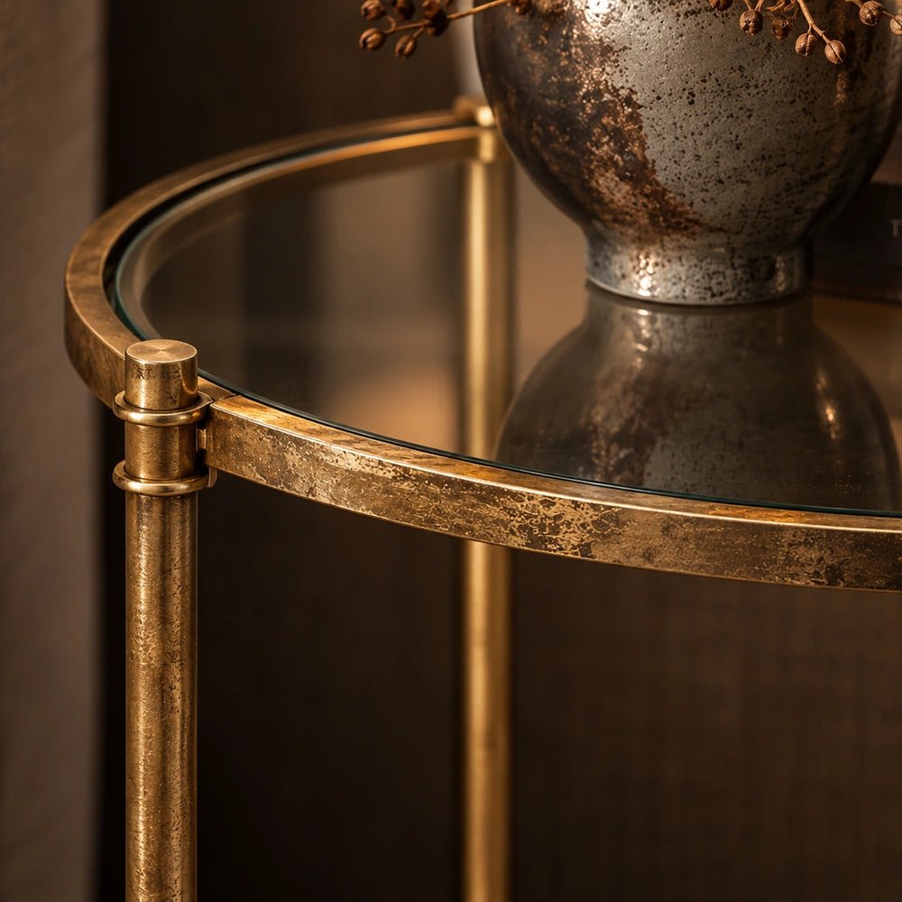 Close detail of the Carson Side Table showing its aged brass frame, circular glass shelf and reflective surface beside a textured vase.