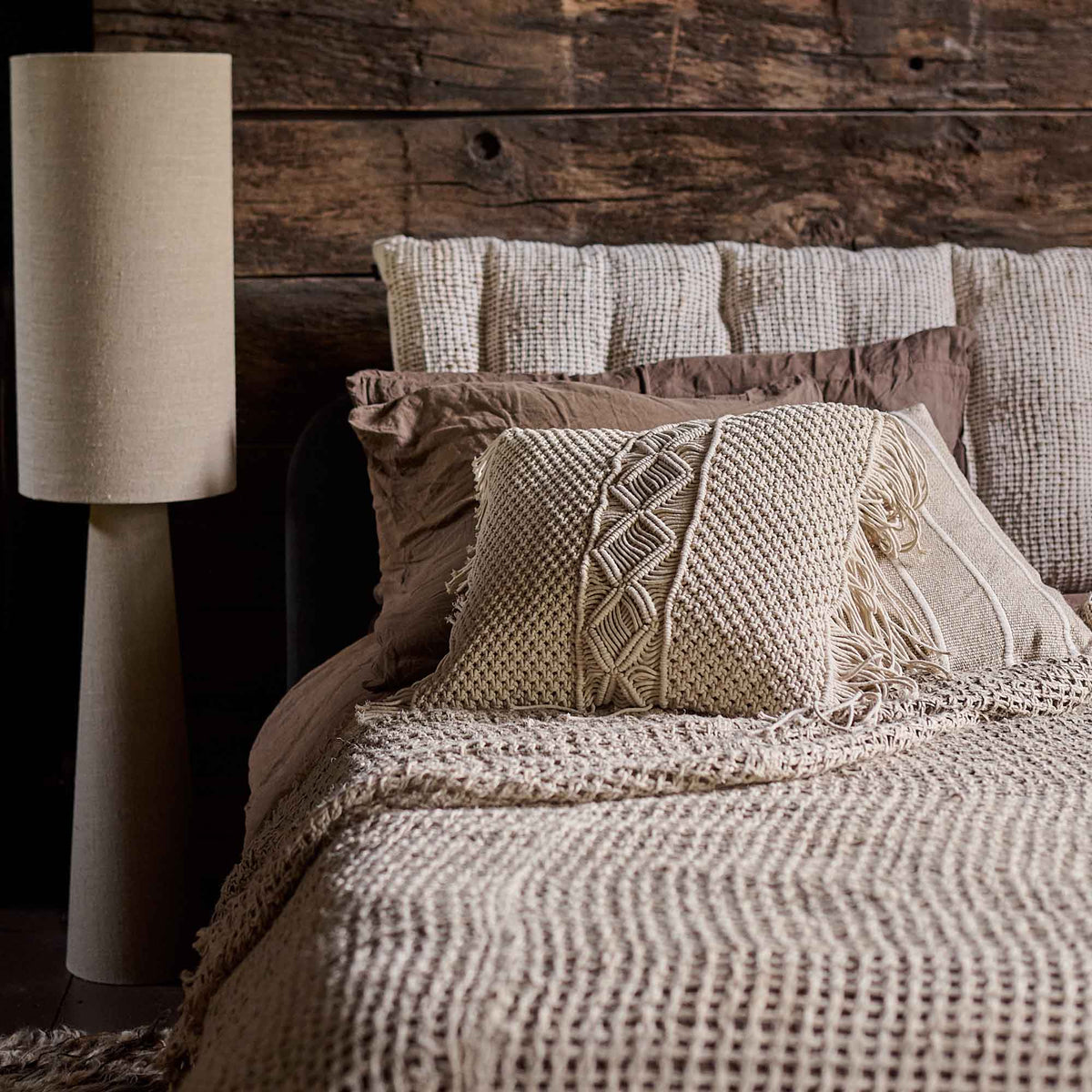 Cassidy Cushion, a square cushion with a cream coloured macrame front and long fringe detail, on a bed next to a tall lamp.