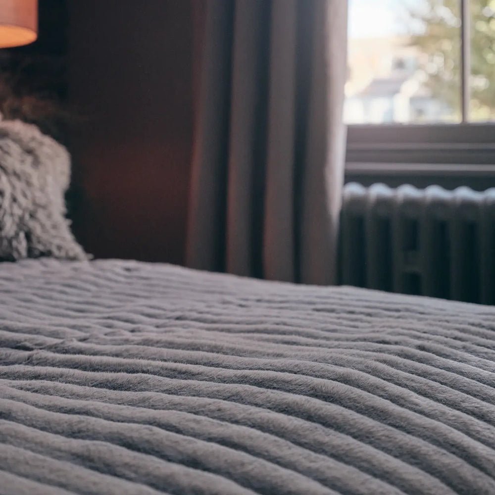 Chandler faux fur throw shown close up across a bed, highlighting its soft taupe colour and raised parallel ribs, with a window in the background.