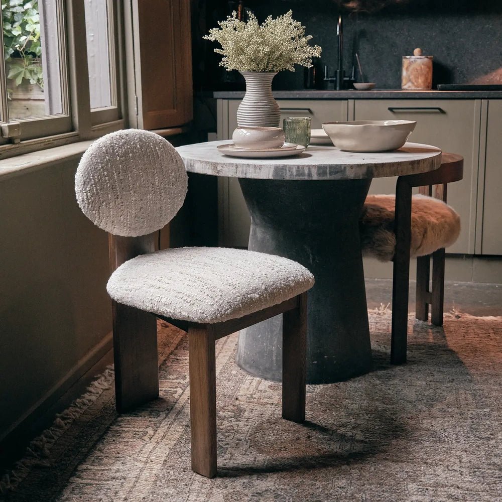 Channing boucle dining chair with a circular cream backrest, textured upholstered seat and warm wooden legs, standing beside a round table in a styled dining room.
