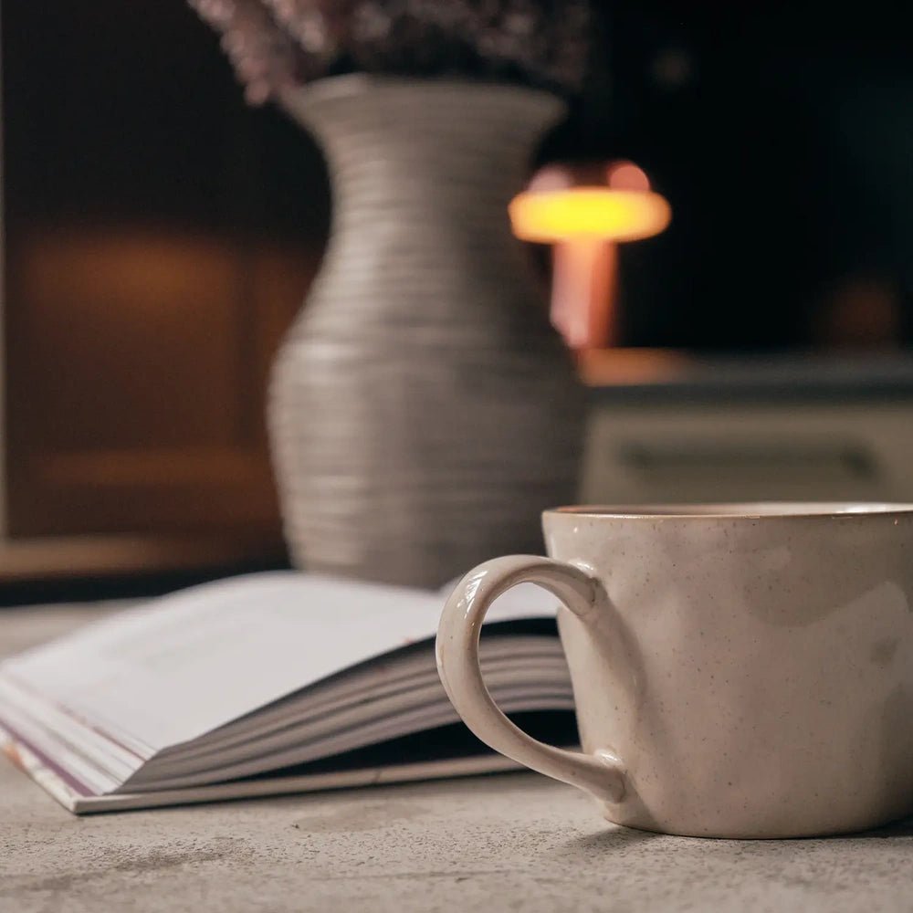 Coleman ceramic mug with a rounded handle and softly mottled cream finish, placed beside an open book on a pale table with a vase behind.