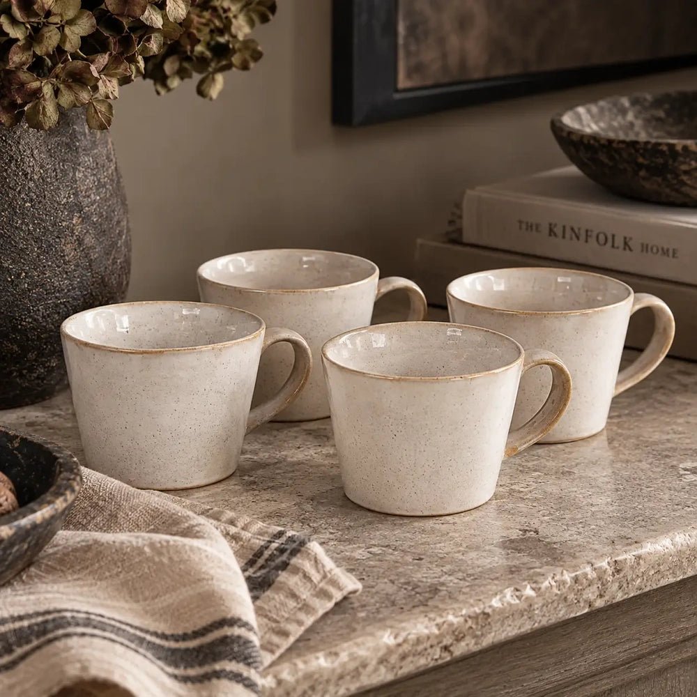 Coleman ceramic mugs in creamy off-white with rounded handles and gently tapered sides, grouped on a stone counter beside striped linen, books and a vase.
