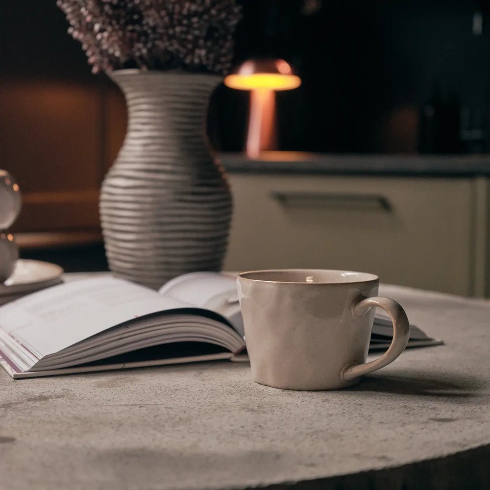 Coleman ceramic mug in creamy beige with a rounded handle, resting on a pale tabletop beside an open book, ribbed vase and softly lit background.