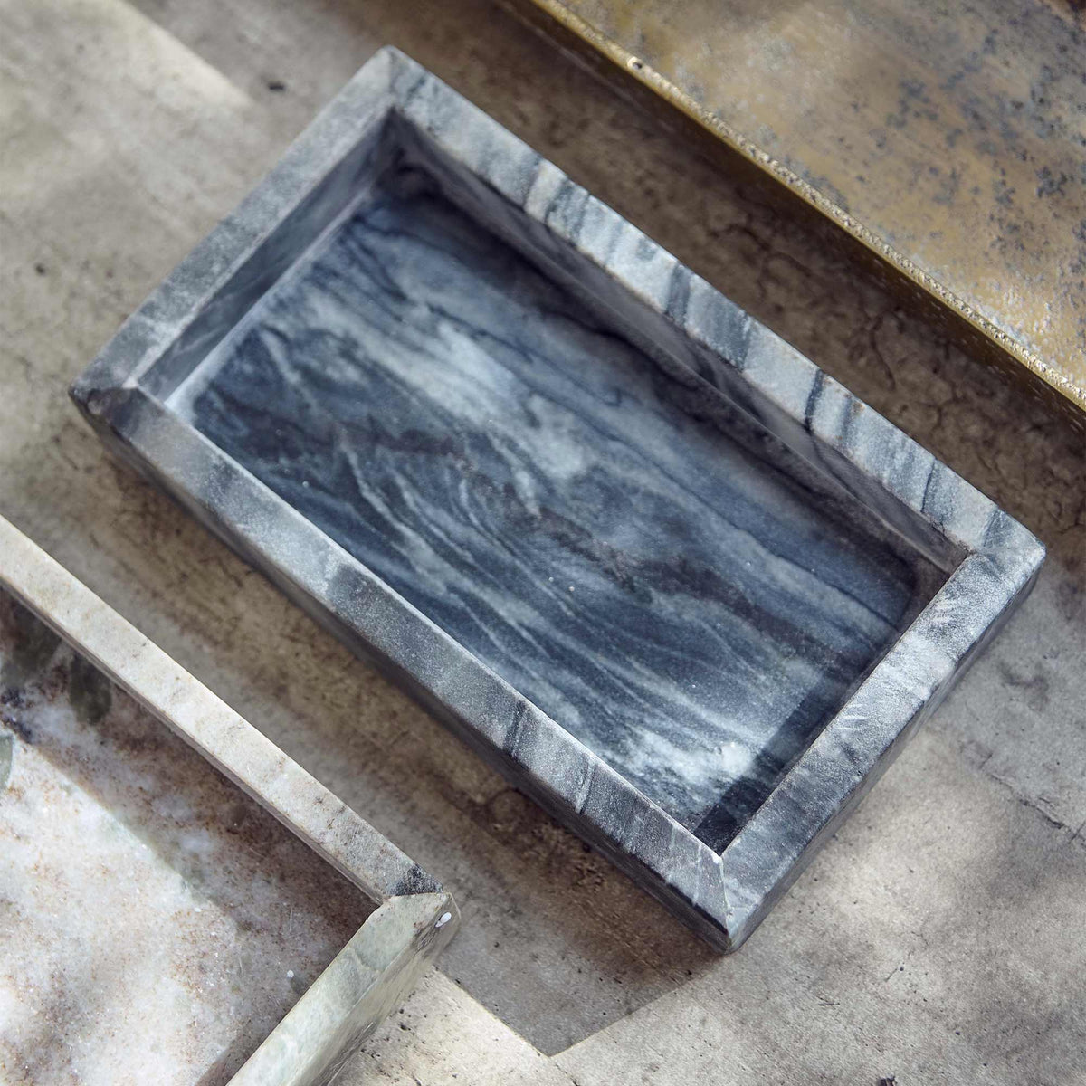 The Creston Marble Tray, a small slim rectangular tray in inky dark hues, next to a selection of decorative trays on a table.