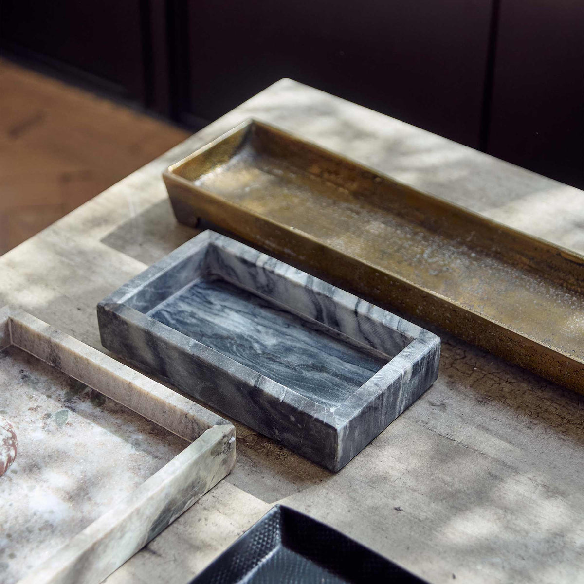 The Creston Marble Tray, a small slim rectangular tray in inky dark hues, next to a selection of decorative trays on a table.