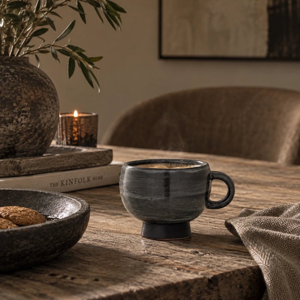 Culross Stoneware Mug with deep charcoal glaze and rounded handle, styled on a pale stone table beside stacked books, candlelight and neutral upholstered seating.