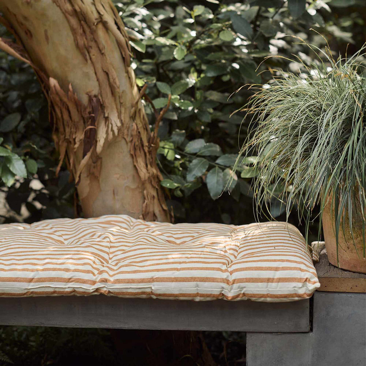 The Daltona Mattress, a slim rectangular mattress in warm ochre and sand coloured stripes, on a concrete bench on a patio.