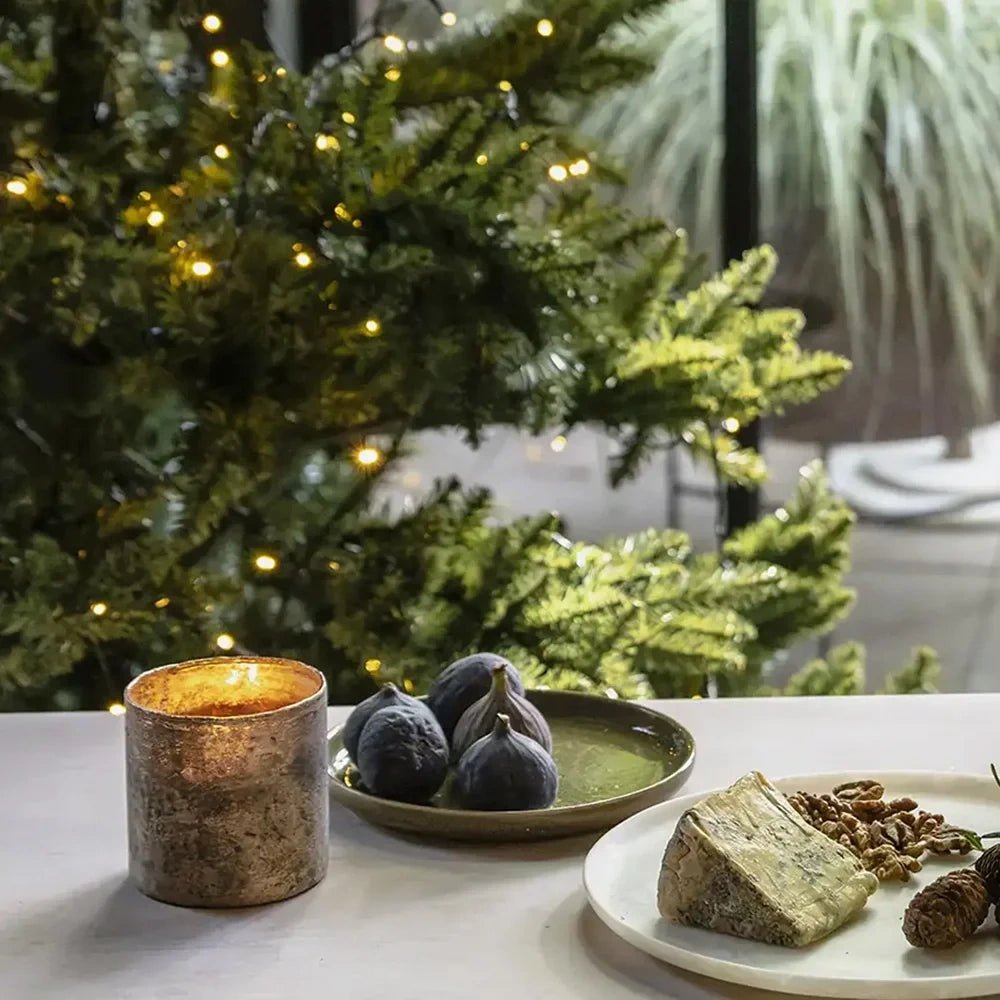 The Dawley glass candle holder has a mottled metallic surface and warm golden glow, standing beside figs and cheese with a lit Christmas tree behind.