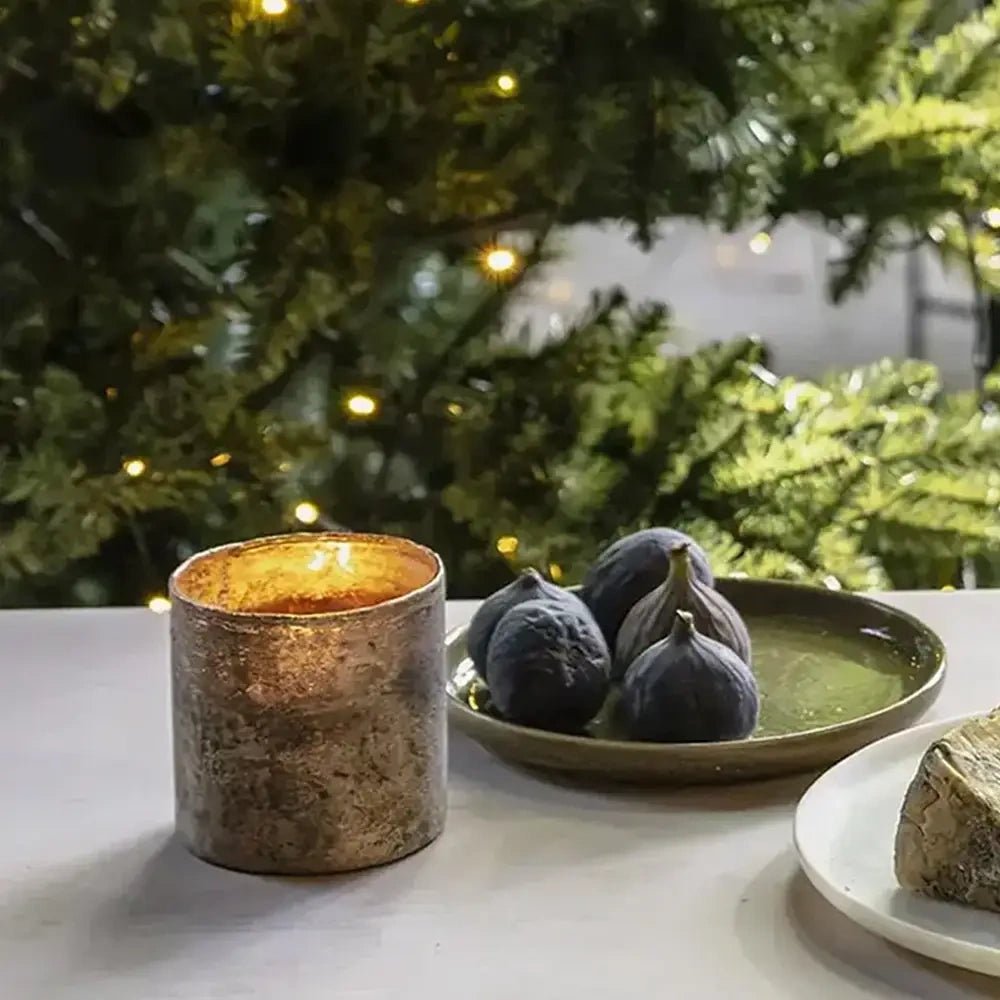 Close-up of the Dawley glass candle holder showing its mottled metallic surface and glowing golden interior beside dark figs, with illuminated Christmas branches behind.