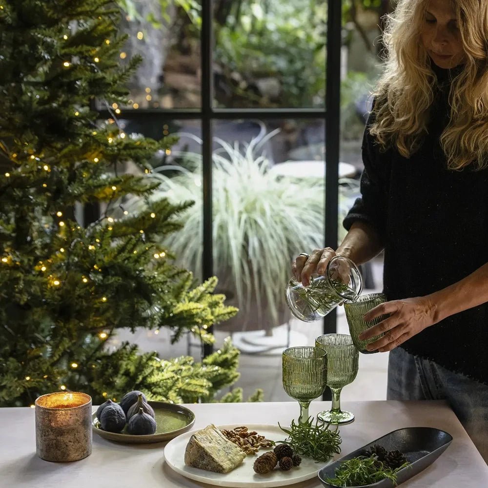 The Dawley glass candle holder glows on a festive table beside figs and cheese while a woman pours a drink into a green ribbed glass.