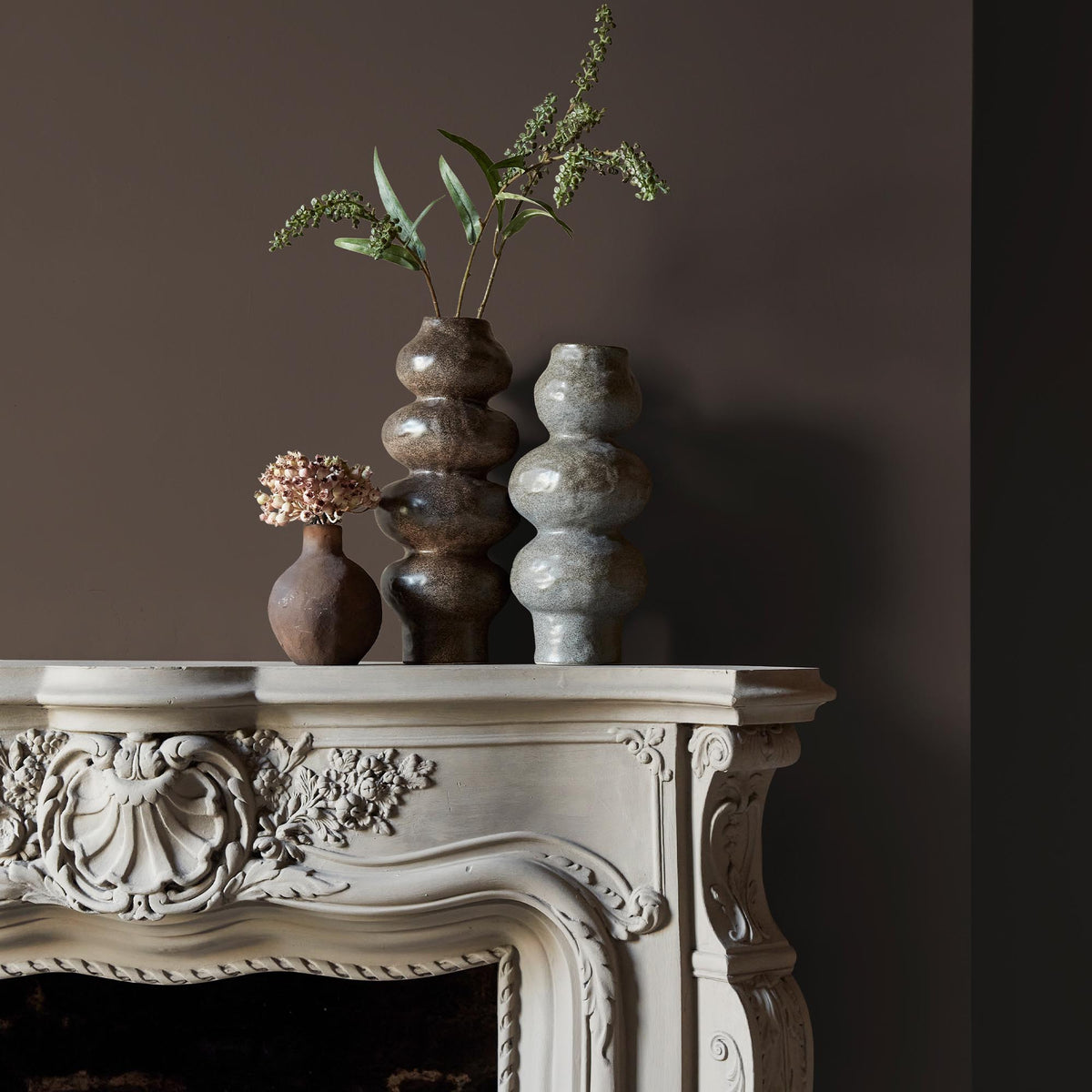 Three ceramic vases sitting on an intricate fireplace mantle in front of a wall in a deep brown paint colour with warm undertones.