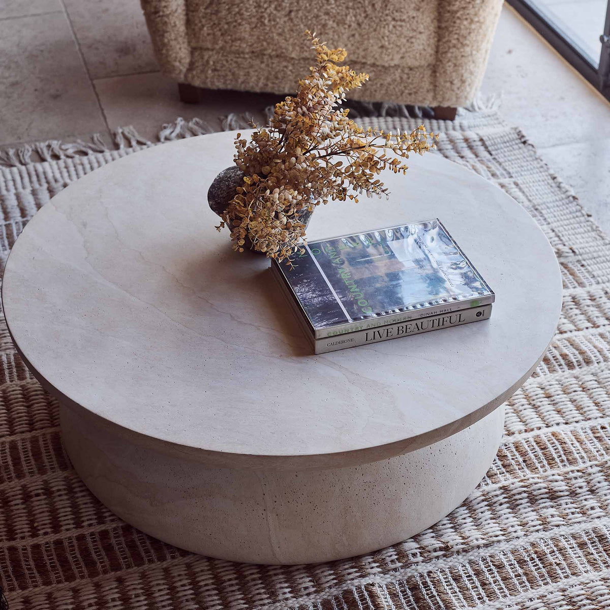 Round travertine effect coffee table with a soft beige finish, subtle cream and grey tones and a sculptural contemporary design. Diablo Round Coffee Table by Abigail Ahern, crafted from glass-reinforced concrete with a textured stone-inspired surface and suitable for both indoor and outdoor spaces. A luxury statement coffee table perfect for modern living rooms and stylish interiors, adding natural texture, timeless elegance and a refined focal point to your home decor.