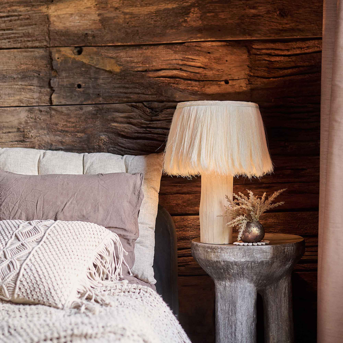 The Dolan Table Lamp, with a natural wooden base and an unusual cream fringed shade made from sisalana fibre, next to a bed.