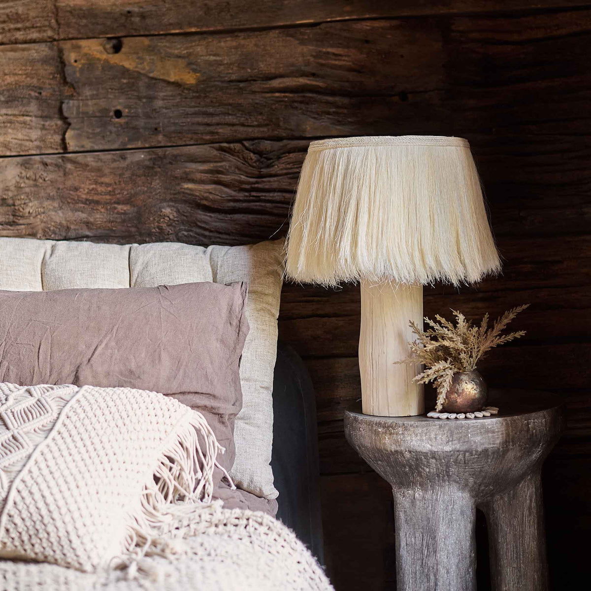 The Dolan Table Lamp, with a natural wooden base and an unusual cream fringed shade made from sisalana fibre, next to a bed.