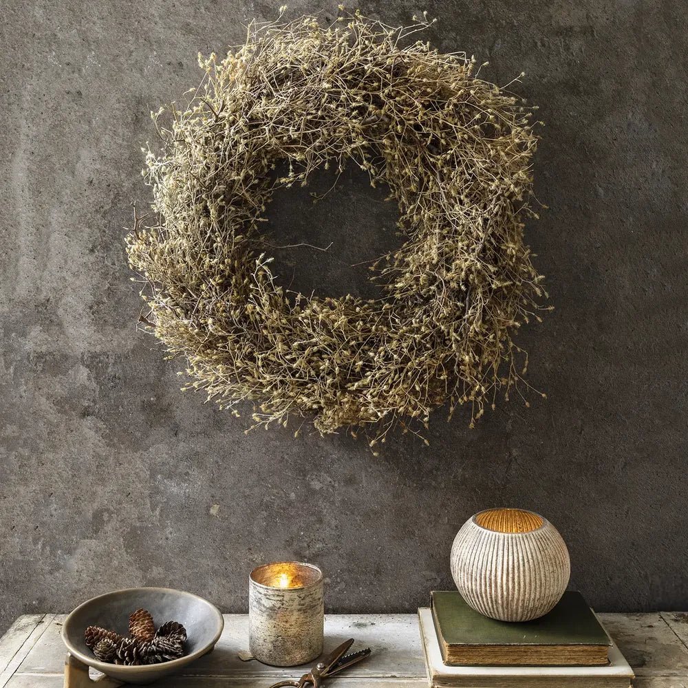 Dried wildwood wreath with an irregular ring of brown foliage hangs on a grey wall above a console styled with a bowl, candle and vase.