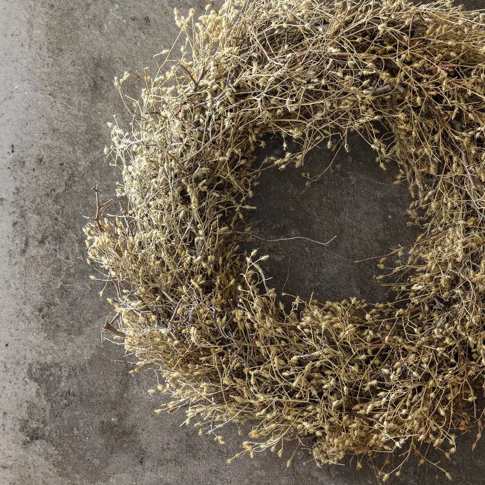Dried wildwood wreath forms a loose circular ring of branching brown foliage against a textured grey wall, with an open centre and uneven outer edge.