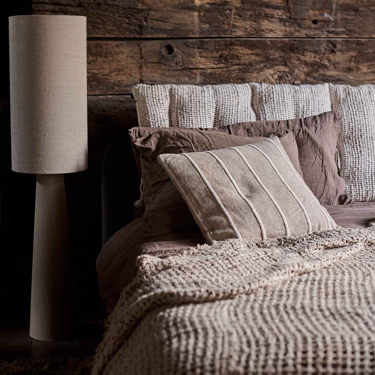 The Edwina Striped Cushion, an elegant sandy beige cushion with ridged piping detail, on a bed next to a tall floor lamp.