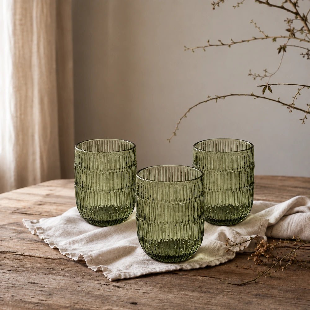Elden Glass Cup, Green with ribbed translucent glass, styled on a moody dining table beside dark plates, cutlery, flowers and layered neutral tableware.