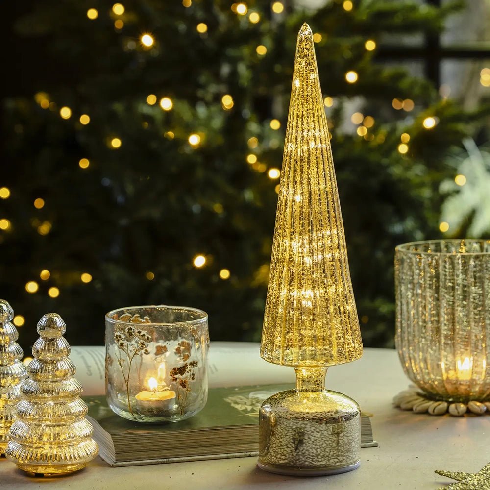 Elira gold tree portable lamp glows through a tall tapered textured cone on a round base, standing beside candles and smaller tree decorations on a festive table.