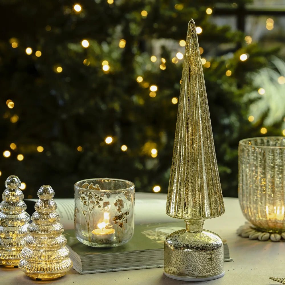 Elira gold tree portable lamp displays an unlit tapered textured cone and rounded base in close view, with a patterned candleholder and Christmas lights behind.