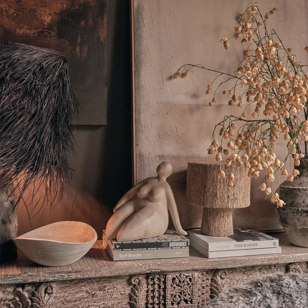 Ellery Figurative Sculpture sits on stacked books within a console arrangement, surrounded by a textured lamp, decorative bowl and tall branches against neutral walls.