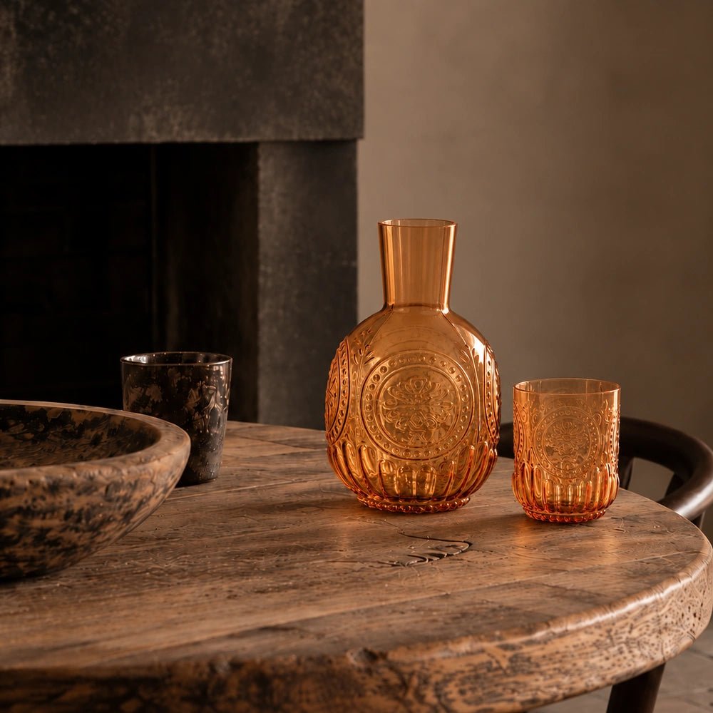Ember Carafe and Cup Set in translucent amber glass, arranged on a rustic wooden table with dark ceramics in a warm softly lit interior.