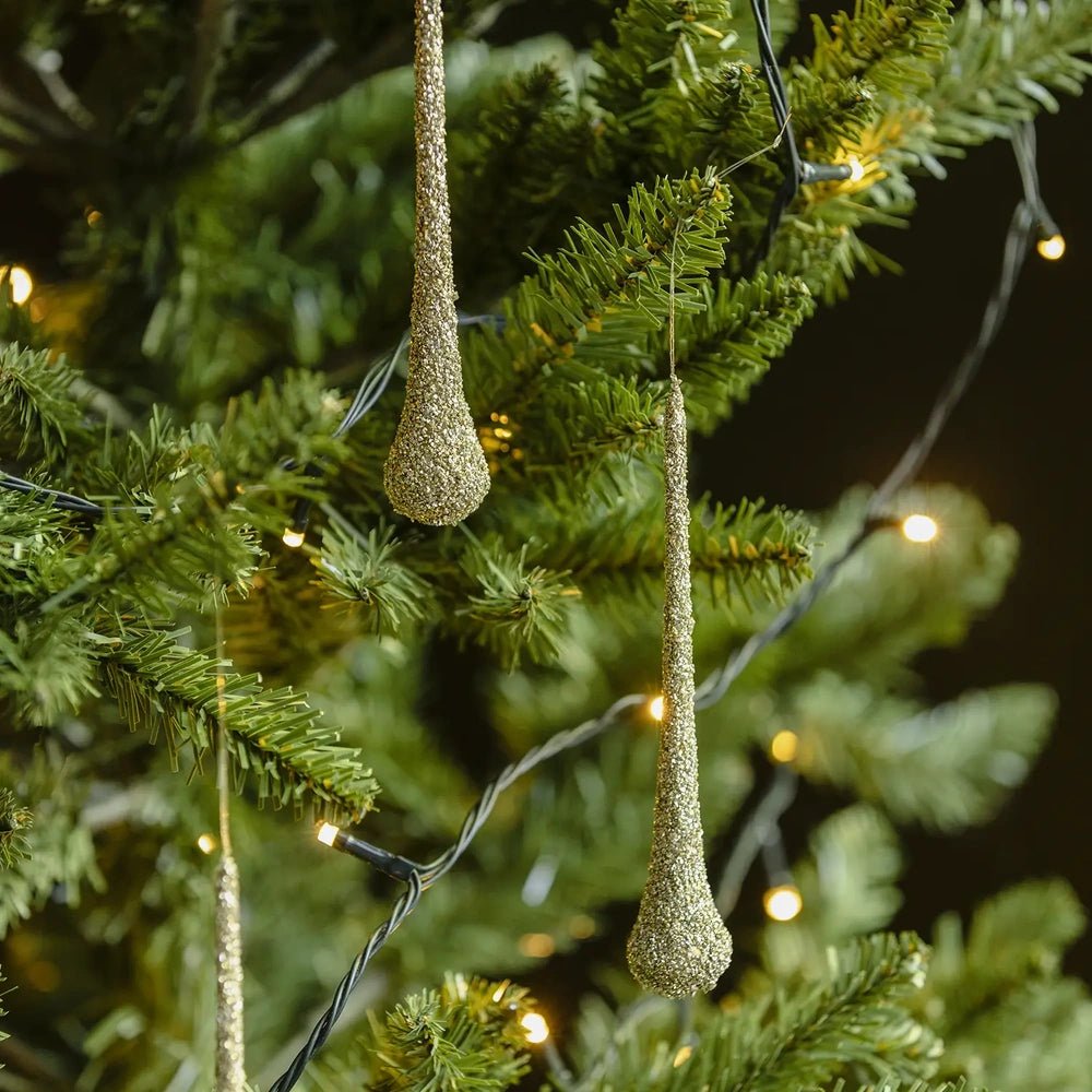 Enzo glitter droplet bauble shows a slender tapered silhouette and sparkling pale gold surface, hanging from a green Christmas tree branch beside warm lights.